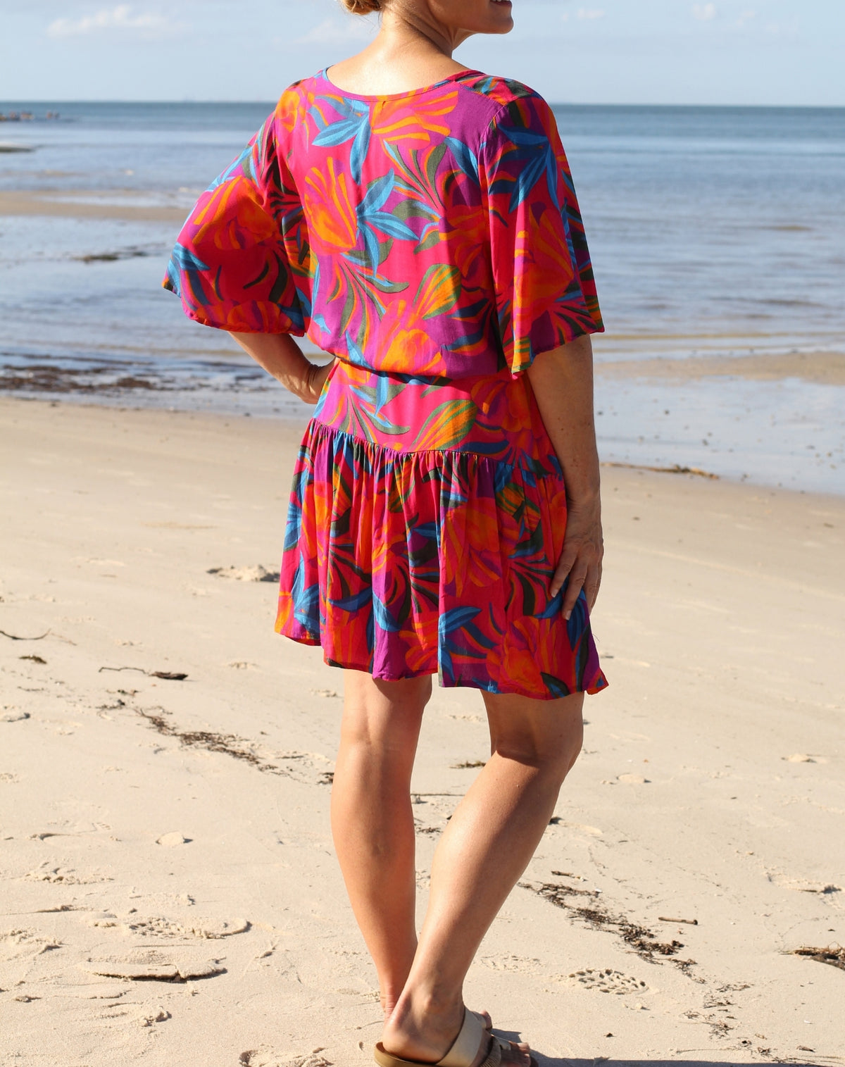 A woman standing on a sandy beach with the ocean in the background, wearing a bright tropical floral print dress with pink, orange, blue, and purple tones.