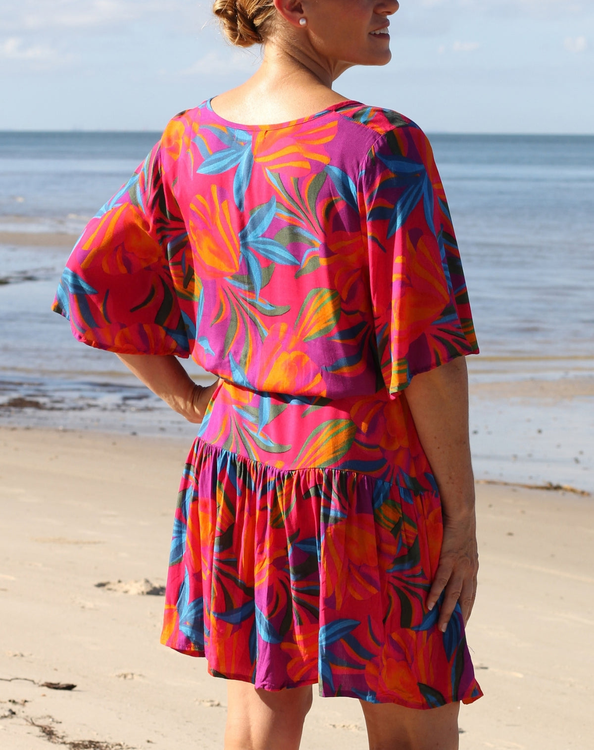 A woman standing on a sandy beach wearing a short boho flutter dress, colorful tropical floral print dress, photographed from the back.