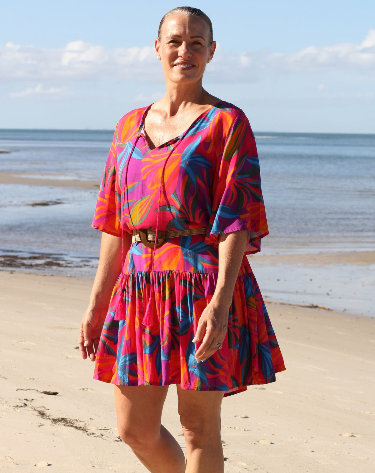 A woman stands on a sandy beach wearing a short, boho flutter dress with a bold tropical floral pattern in pink, orange, blue, and purple, cinched at the waist with a brown belt.