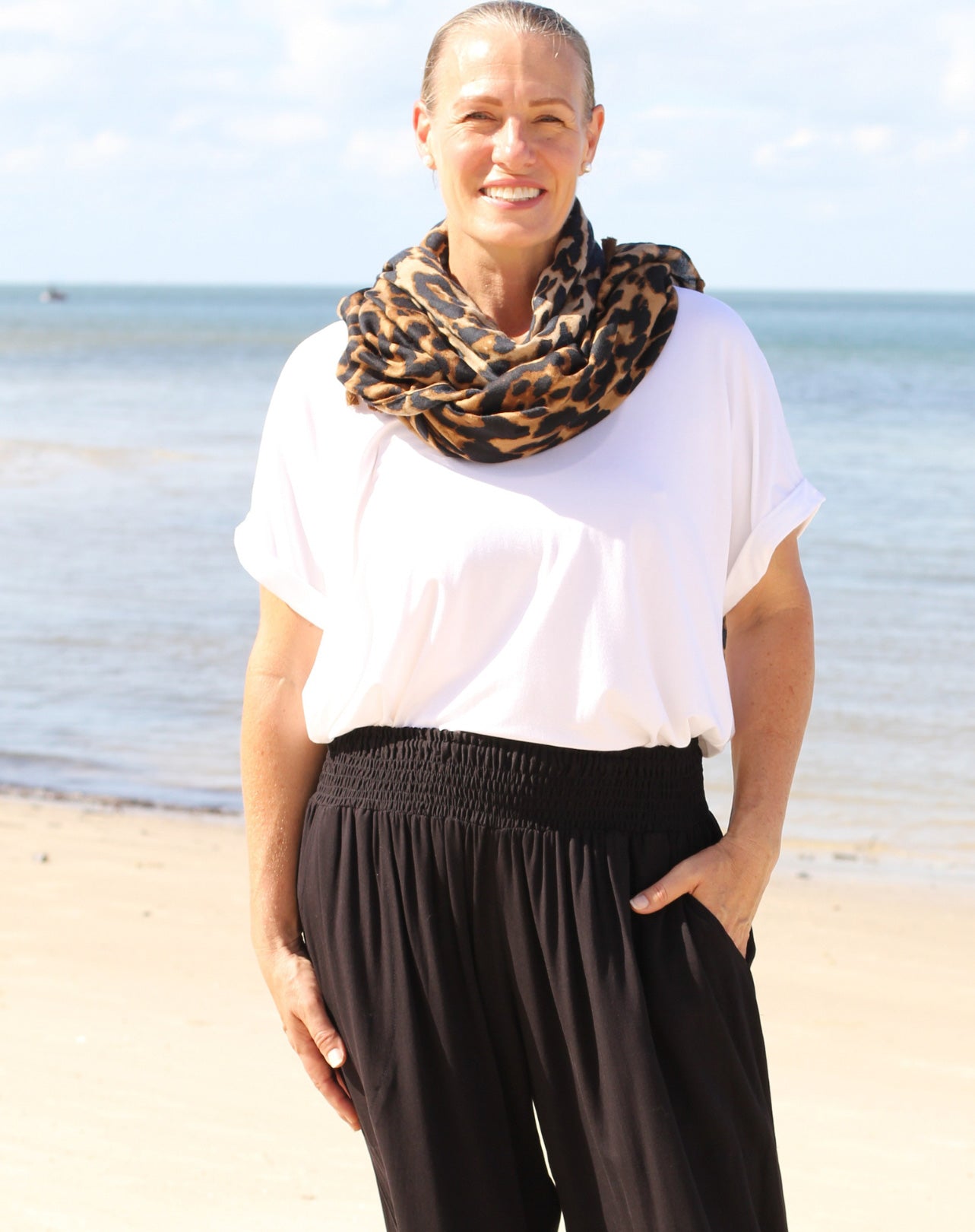 Woman standing on a beach wearing a white modal tee top and black pants with a leopard print scarf.
