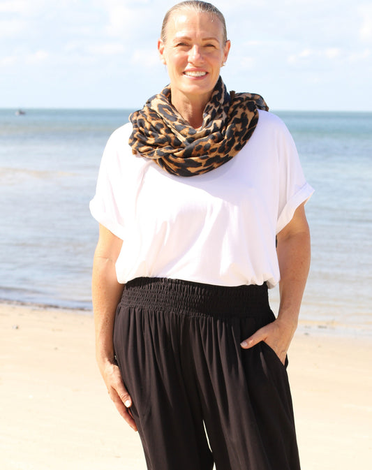Woman standing on a beach wearing a white modal tee top and black pants with a leopard print scarf.