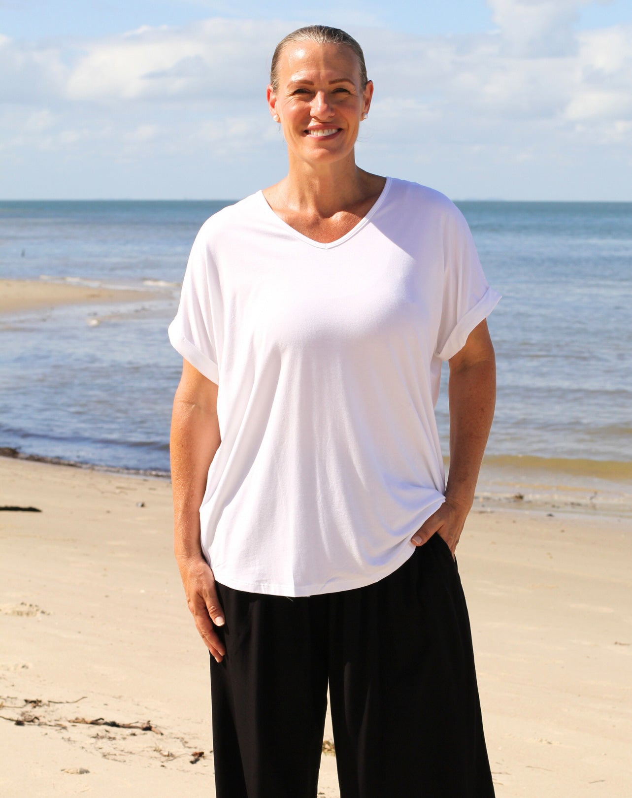 Person wearing a white classic v t-shirt and black pants standing on a beach with ocean and sky in the background