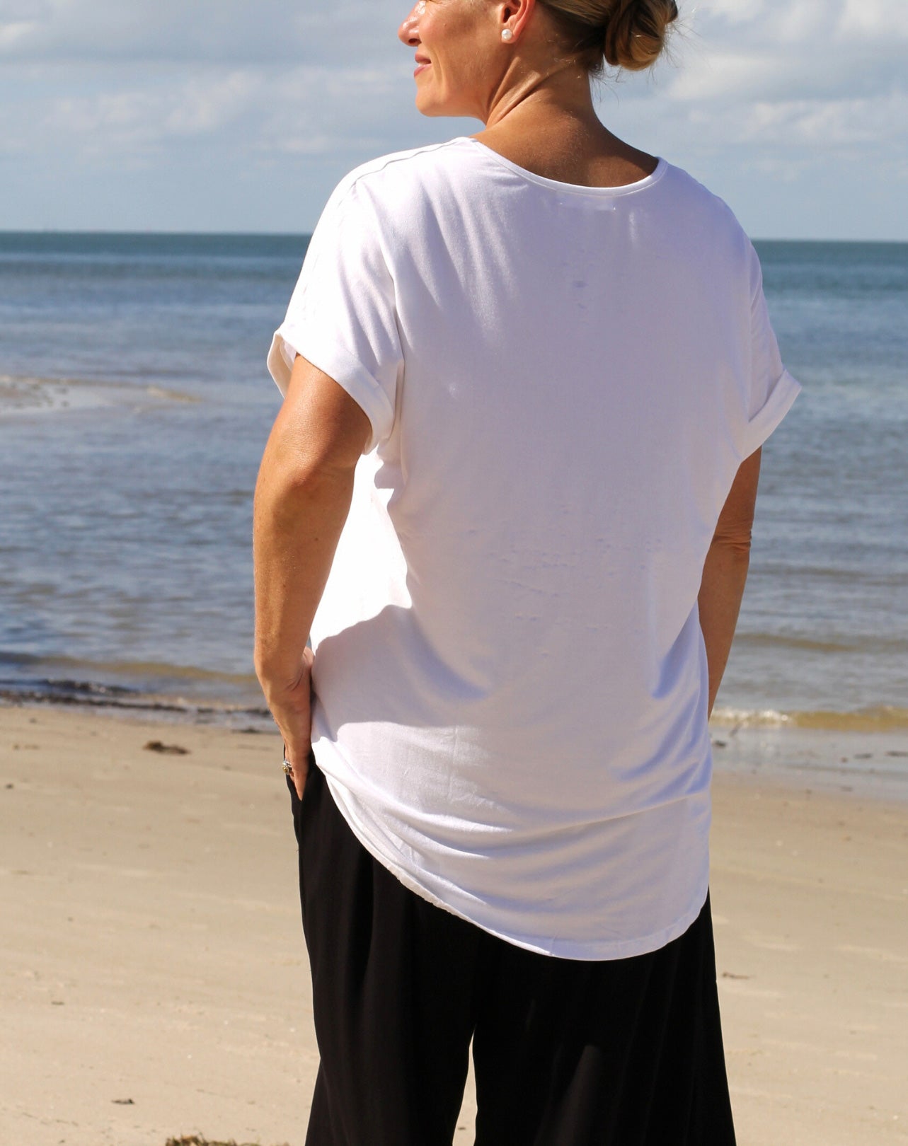 Person wearing a white classic v t-shirt and black pants standing on a beach with ocean in the background