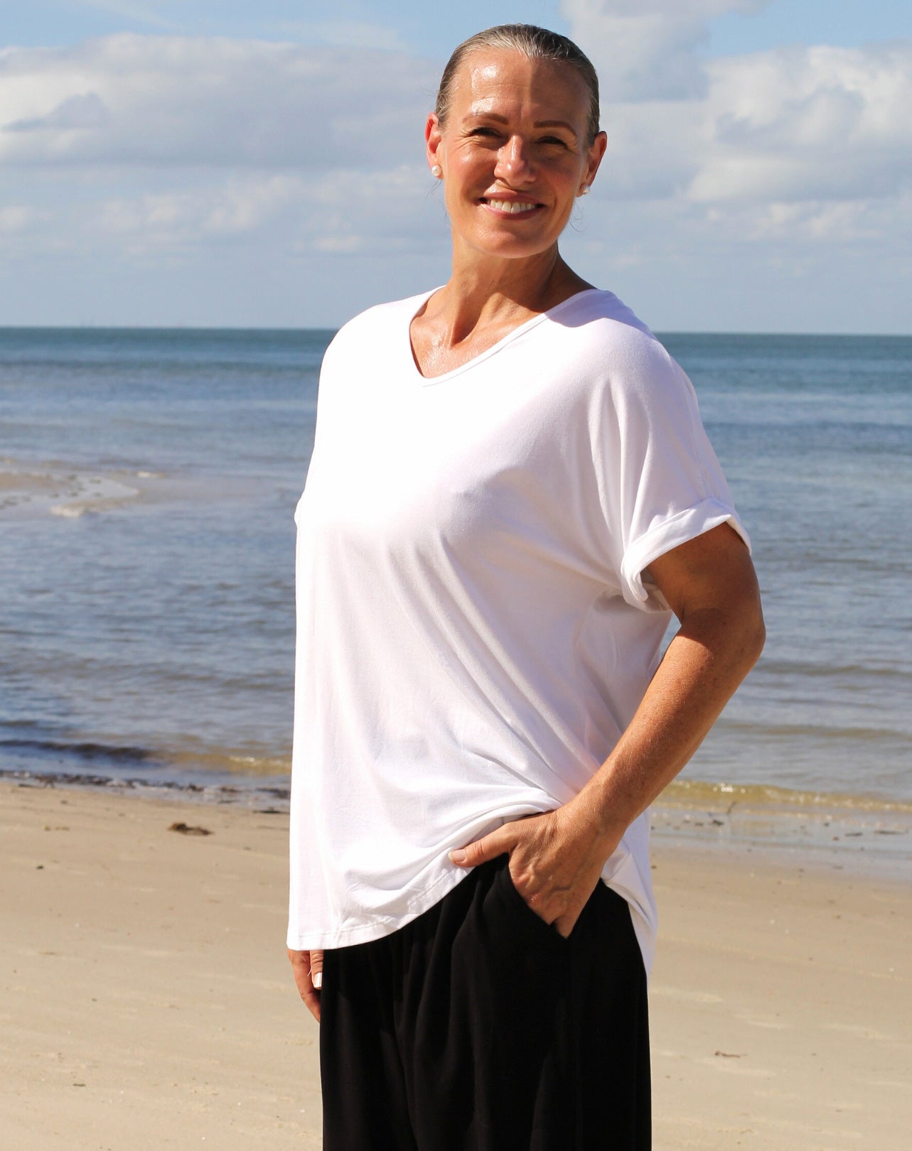 Person wearing a white v neck modal tee shirt and black pants standing on a beach with ocean and sky in the background