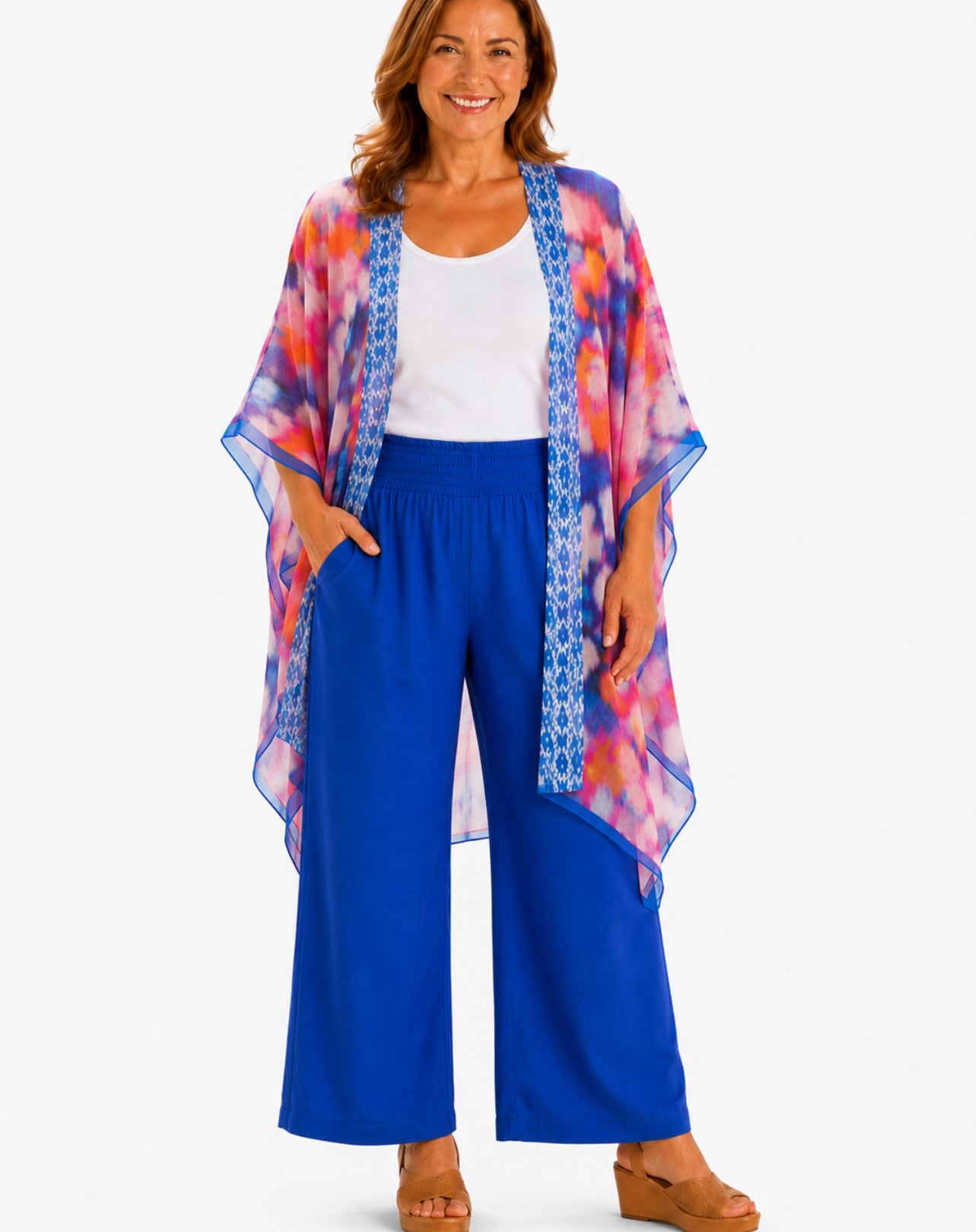 A woman modeling a lightweight open-front kimono cardigan with a vibrant blue, pink, and orange tie-dye pattern and blue patterned trim, worn over a white tank top and bright blue wide-leg pants.