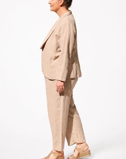 A woman shown in side profile wearing a light beige linen two-piece set consisting of a cropped blazer and wide-leg trousers, paired with beige wedge sandals against a plain white background.