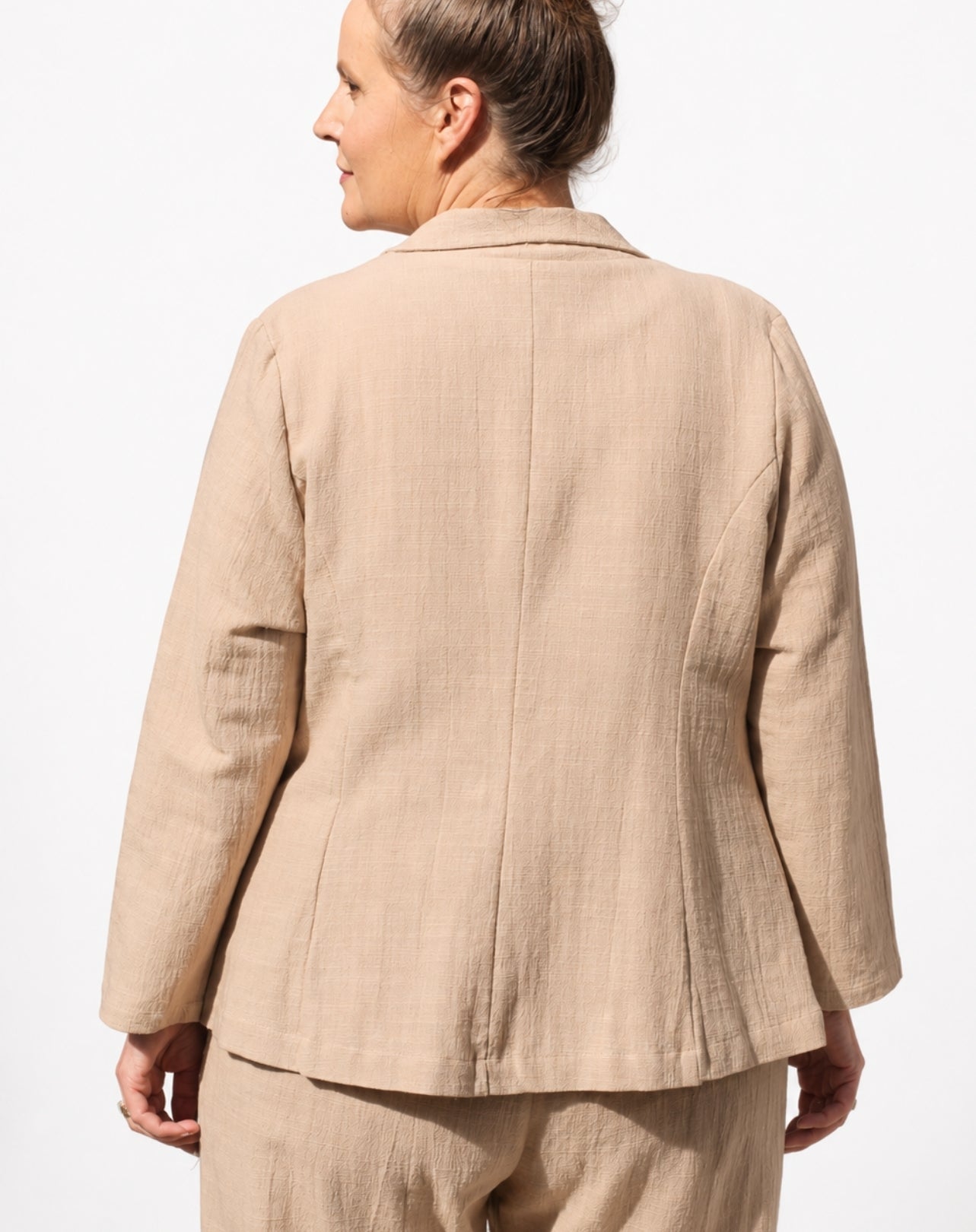 A woman shown from the back wearing a light beige linen blazer and matching trousers against a plain white background.