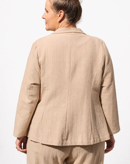 A woman shown from the back wearing a light beige linen blazer and matching trousers against a plain white background.