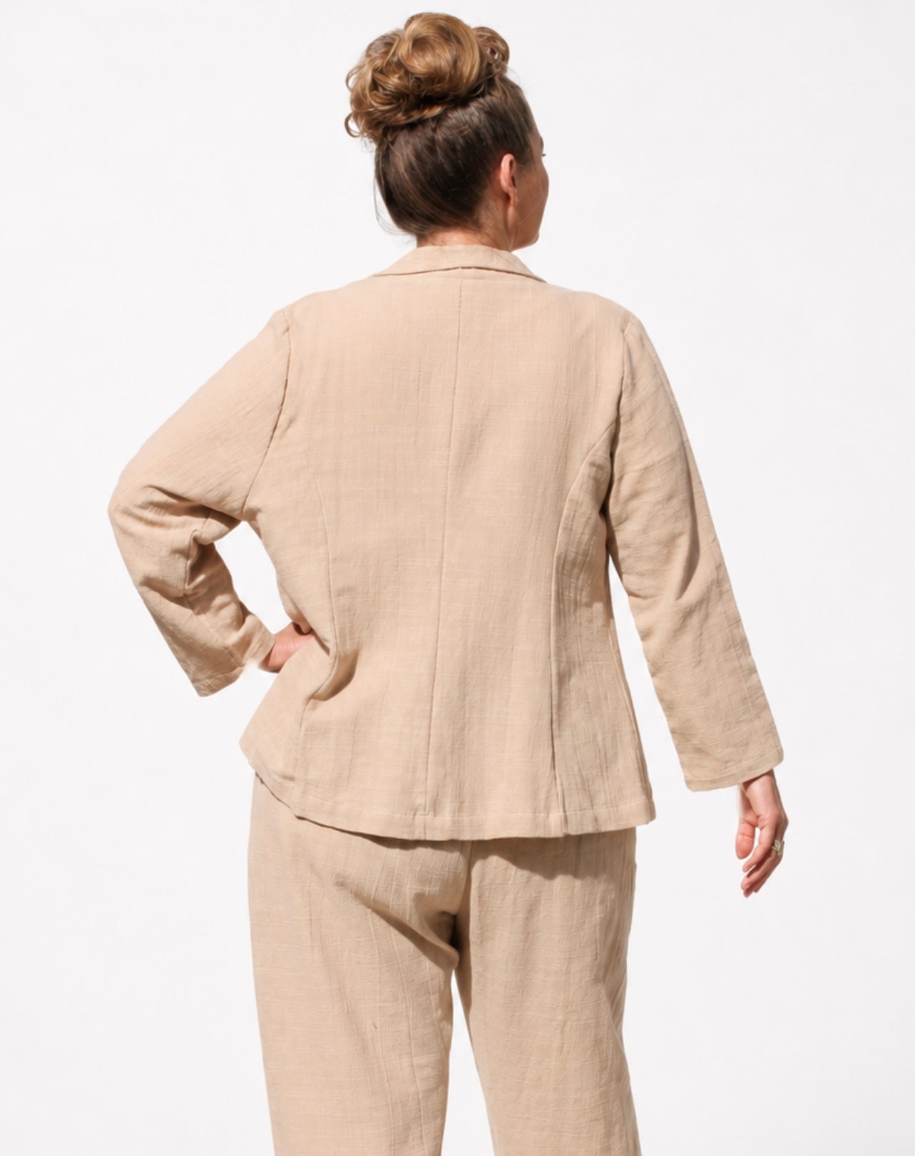 A woman shown from the back wearing a light beige linen two-piece suit, consisting of a long-sleeve blazer and matching trousers, against a plain white background.