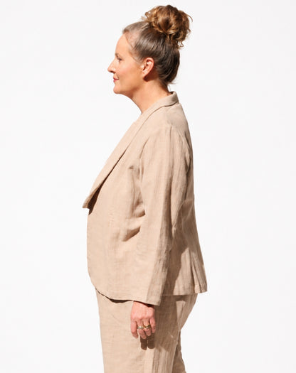 A woman shown in side profile wearing a light beige linen suit, featuring a cropped blazer and matching trousers, against a plain white background.