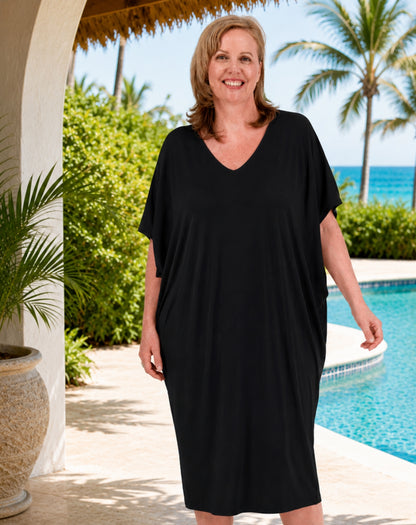 A woman wearing a comfy black short-sleeve V-neck cover-up dress stands outdoors by a swimming pool with palm trees and blue water in the background.
