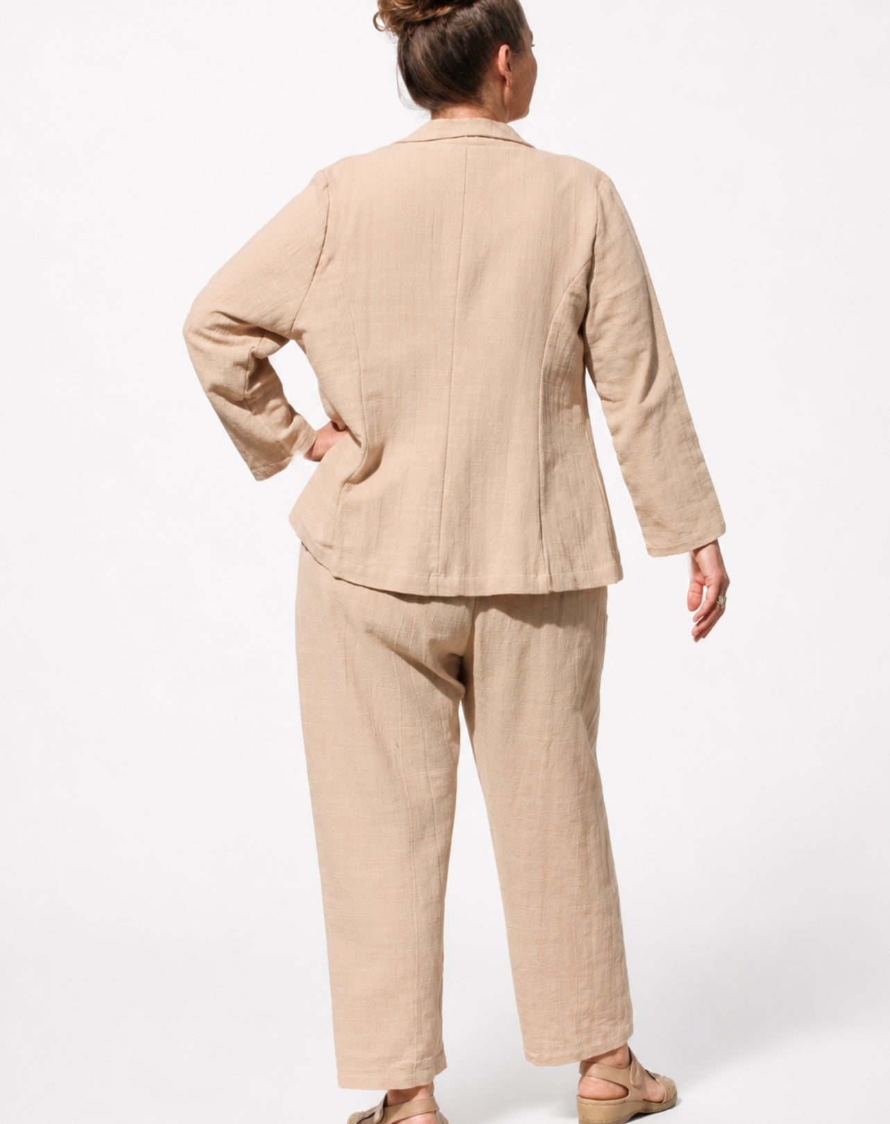 A woman shown from the back wearing a light natural linen two-piece set consisting of a long-sleeve blazer jacket and matching relaxed leg trousers, photographed against a clean white studio background.