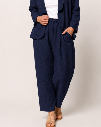A person wearing a navy blue linen-blend two-piece set consisting of a cropped blazer and wide-leg trousers, styled with a white top and beige sandals against a light gray studio background.