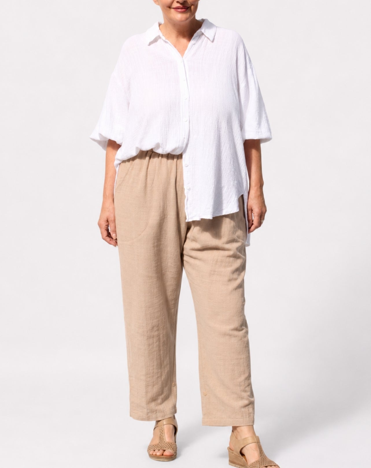 A woman modeling a white short-sleeve button-up shirt and beige linen trousers with tan sandals, photographed against a clean white studio background.