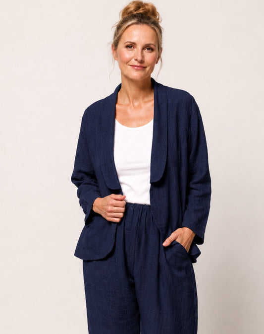A woman modelling a navy blue linen-blend two-piece set consisting of a draped open-front blazer jacket and matching trousers, worn over a white tank top, against a plain light beige studio background.