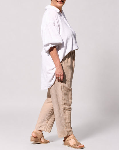 A woman modelling a white oversized short-sleeve blouse and beige natural linen wide-leg pants with tan wedge sandals, shown in profile against a light grey studio background.