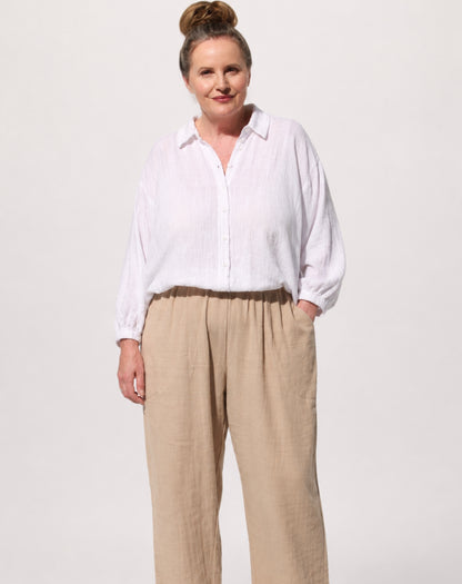 A woman modeling a white comfy cotton long-sleeve button-up shirt and beige wide-leg linen pants against a plain light gray studio background.