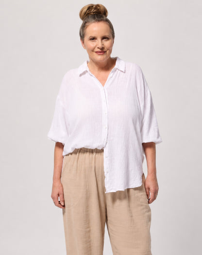 A woman modeling a white long-sleeve button-up shirt with elasticised sleeves and beige wide-leg trousers against a light gray studio background.
