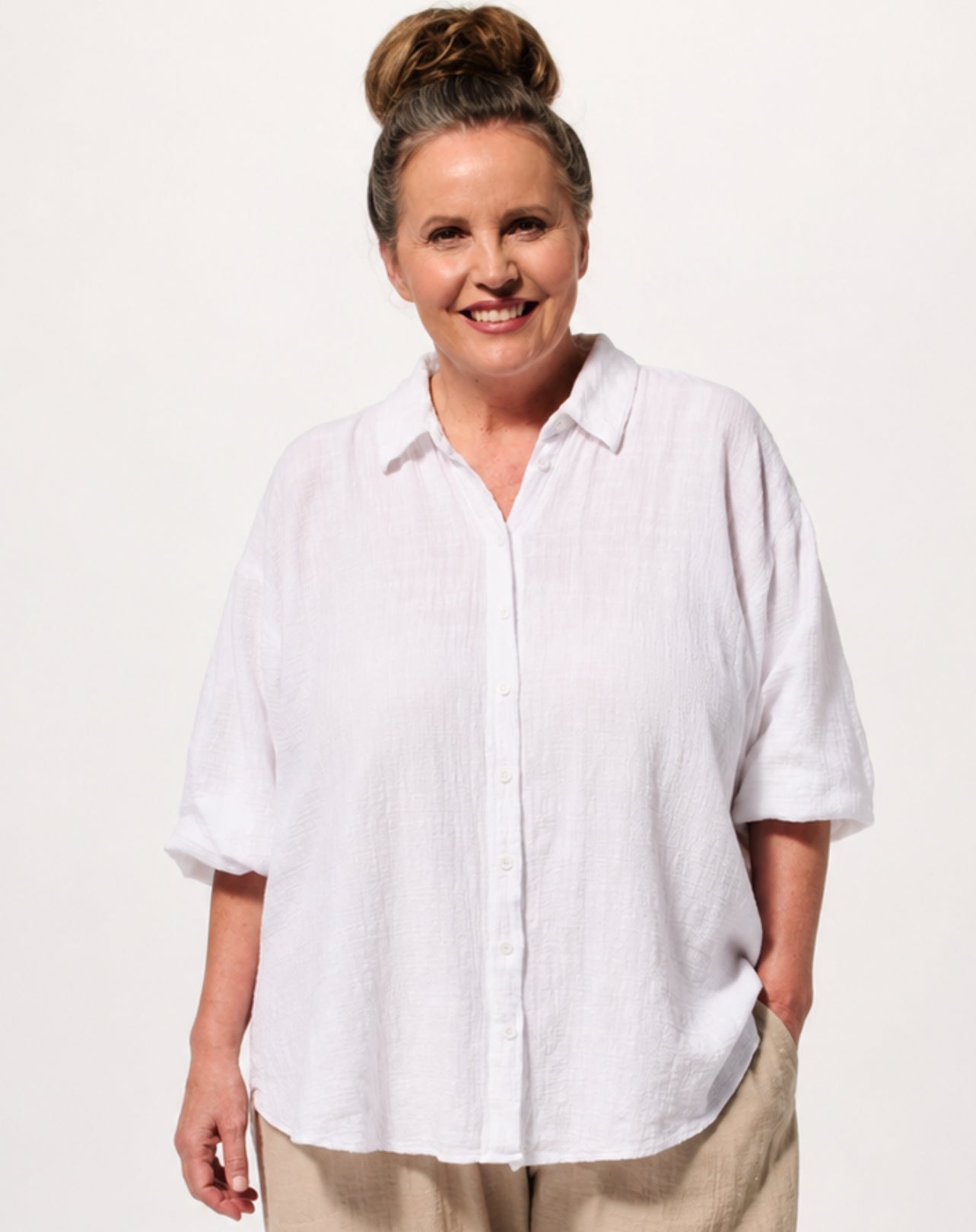 A smiling woman with her hair in a bun wears an oversized white short-sleeve button-up beach shirt and beige pants against a plain light background.