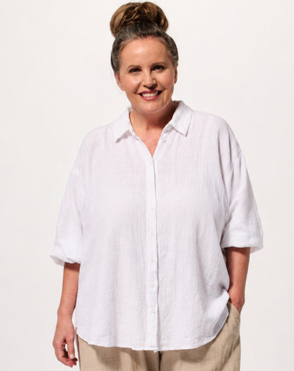A smiling woman with her hair in a bun wears an oversized white short-sleeve button-up beach shirt and beige pants against a plain light background.