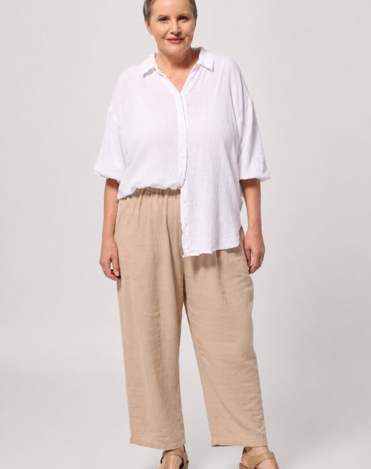 A woman modelling a white comfort cotton shirt adjustable sleeves button-up shirt and beige wide-leg pants against a light grey studio background.