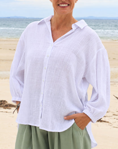 A woman wearing a white long-sleeve white cotton linen look button-up shirt and sage green pants, standing on a sandy beach with the ocean in the background.