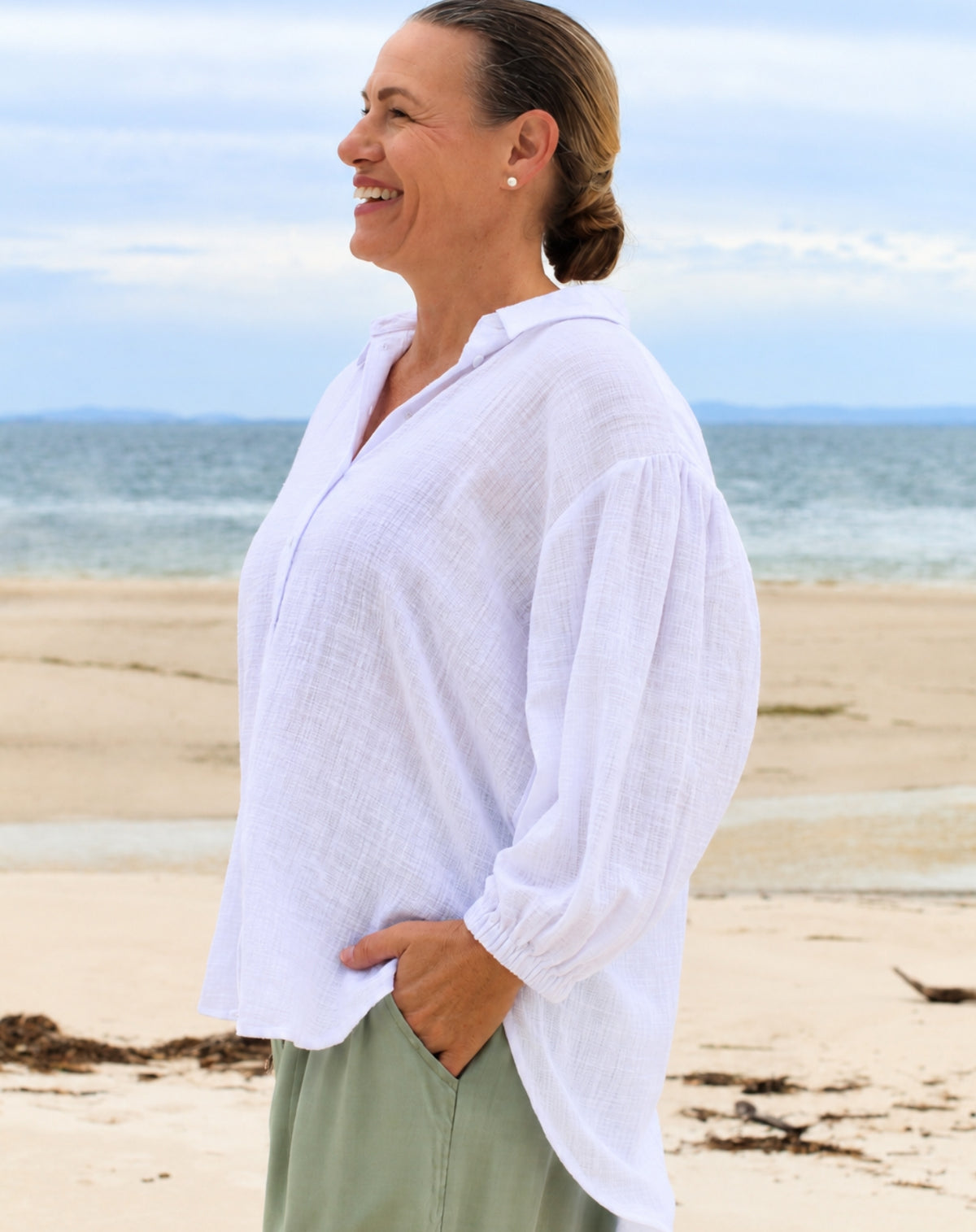 A woman standing on a sandy beach wearing a loose white long-sleeve cotton button-up shirt and sage green pants, with the ocean and cloudy sky in the background.