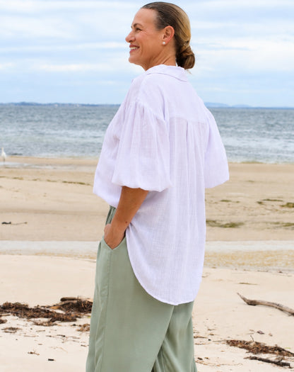 A woman standing on a sandy beach wearing a loose white short-sleeve button-up shirt and sage green wide-leg pants, with the ocean and cloudy sky in the background.