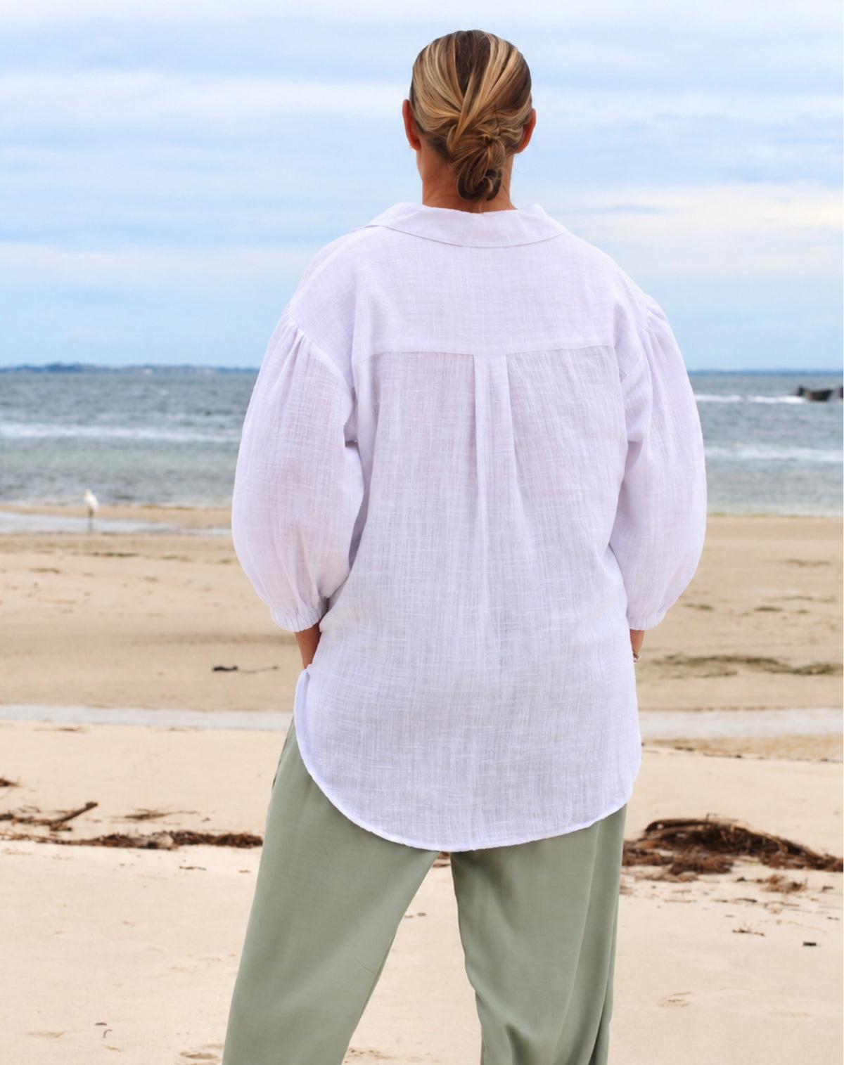 A woman standing on a sandy beach with the ocean and cloudy sky in the background, viewed from behind. She is wearing a loose white linen button-up shirt with voluminous sleeves and pleated back detail, paired with light olive green pants.