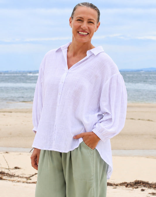 A woman standing on a sandy beach wearing a white long-sleeve button-up shirt and sage green pants.