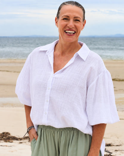 A smiling woman standing on a sandy beach wearing a loose white long-sleeve button-up shirt with adjustable elasticized sleeve and sage green pants, with the ocean and sky in the background.