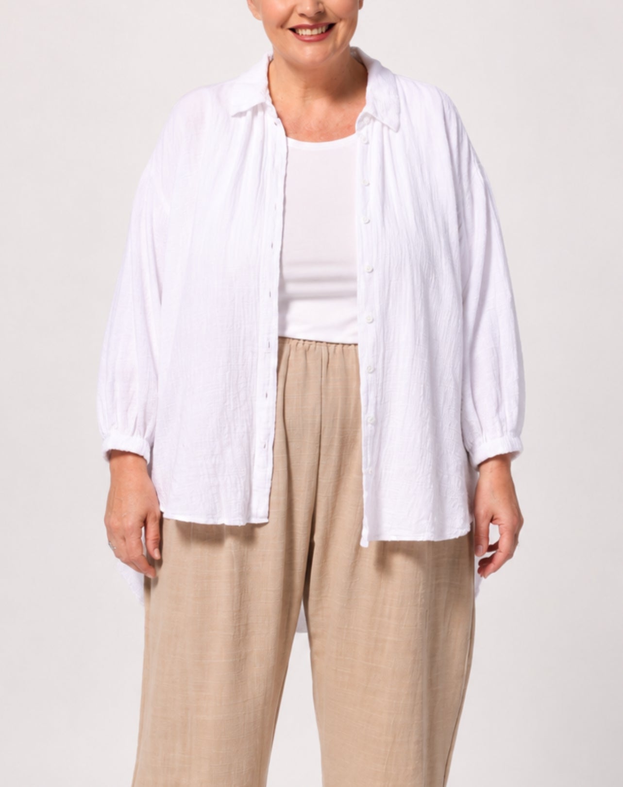 A woman wearing a white long-sleeve button-up shirt over a white tank top and beige wide-leg pants, photographed against a light gray studio background.