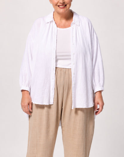 A woman wearing a white long-sleeve button-up shirt over a white tank top and beige wide-leg pants, photographed against a light gray studio background.
