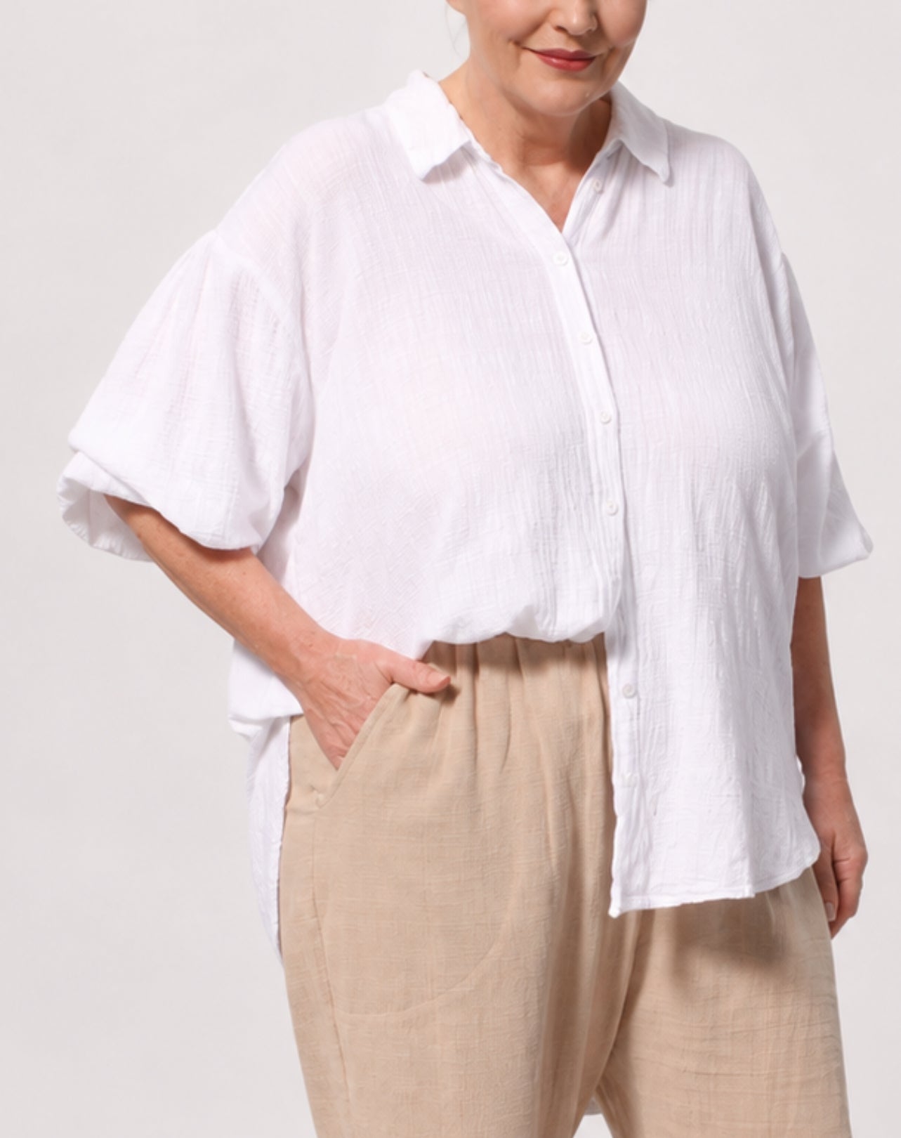 A woman wearing a white adjustable sleeve button-up shirt with a relaxed, oversized fit and beige linen trousers, shown from the chest down against a light grey studio background.