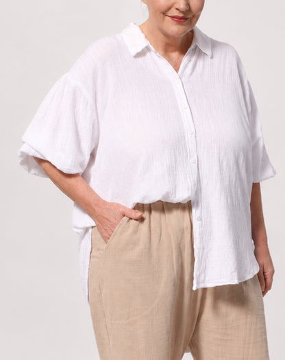 A woman wearing a white adjustable sleeve button-up shirt with a relaxed, oversized fit and beige linen trousers, shown from the chest down against a light grey studio background.