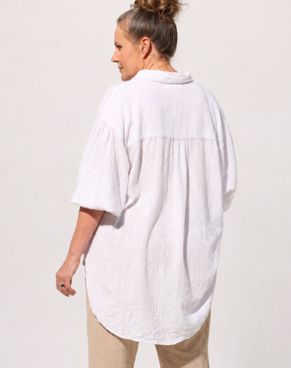 A woman shown from the back wearing a loose oversized cotton white linen look shirt with gathered detailing at the shoulders and waist, paired with beige pants, against a plain light background.