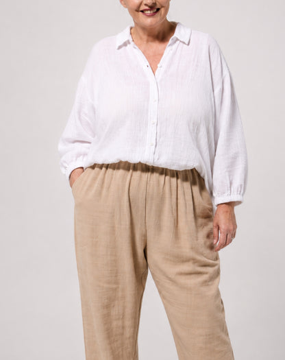 A woman wearing a white long-sleeve cotton button-up shirt and beige linen trousers, standing against a light gray studio background.
