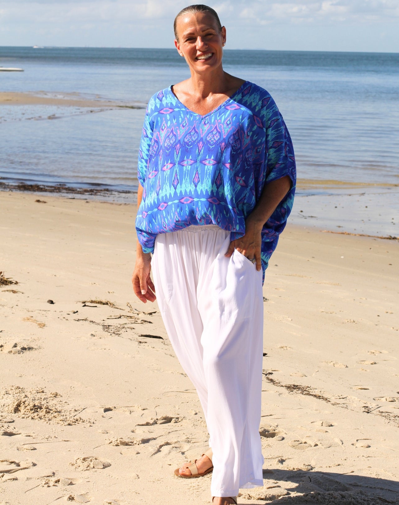 A woman standing on a sandy beach wearing a loose blue patterned top and white pants, with the ocean and sky in the background.