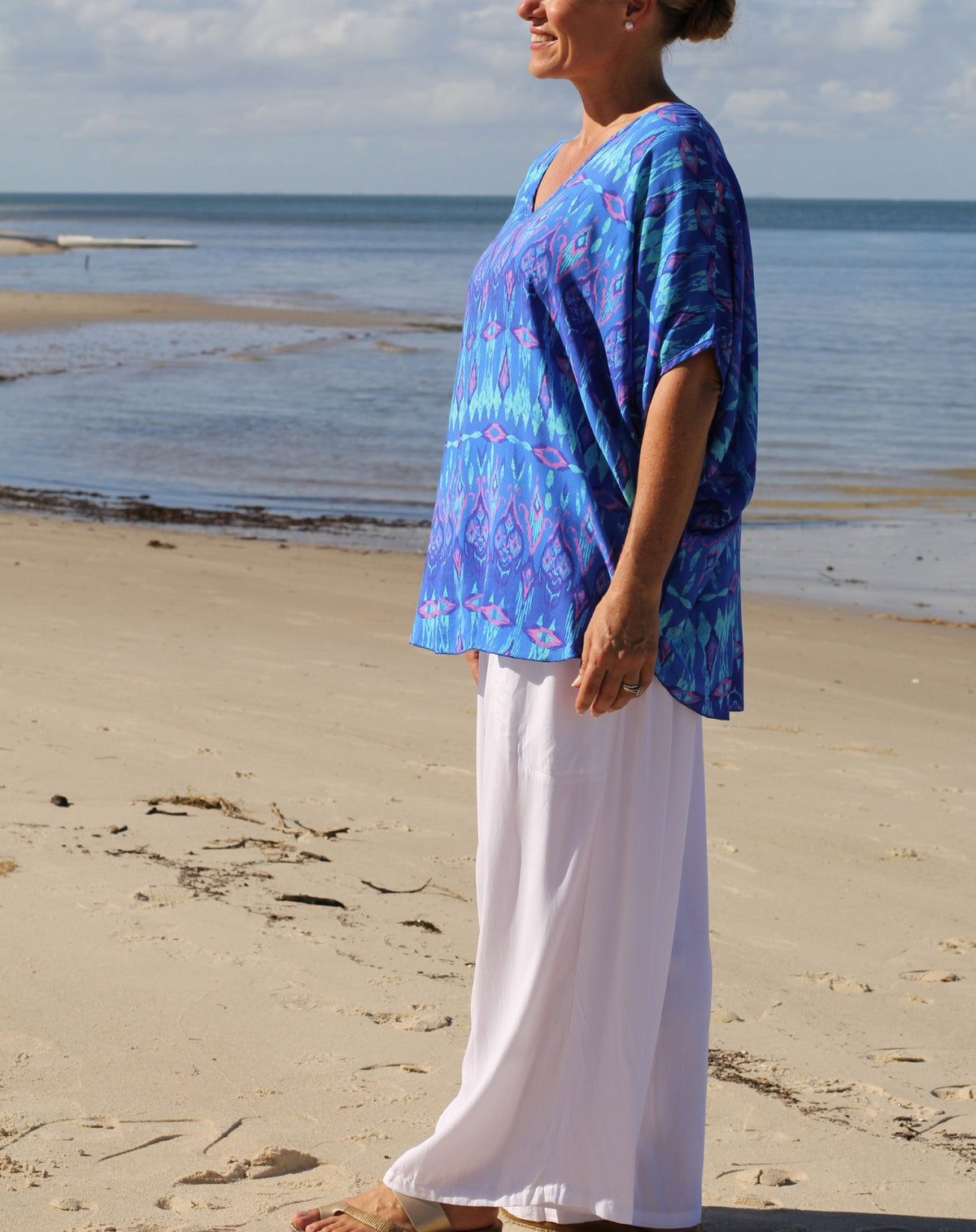 A woman standing on a sandy beach wearing a loose blue patterned tunic top and white wide-leg pants, with the ocean and sky in the background.