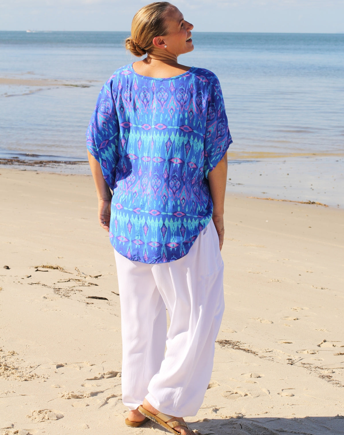 A woman standing on a sandy beach facing the ocean, wearing a loose blue and purple patterned tunic top with white wide-leg pants and sandals.