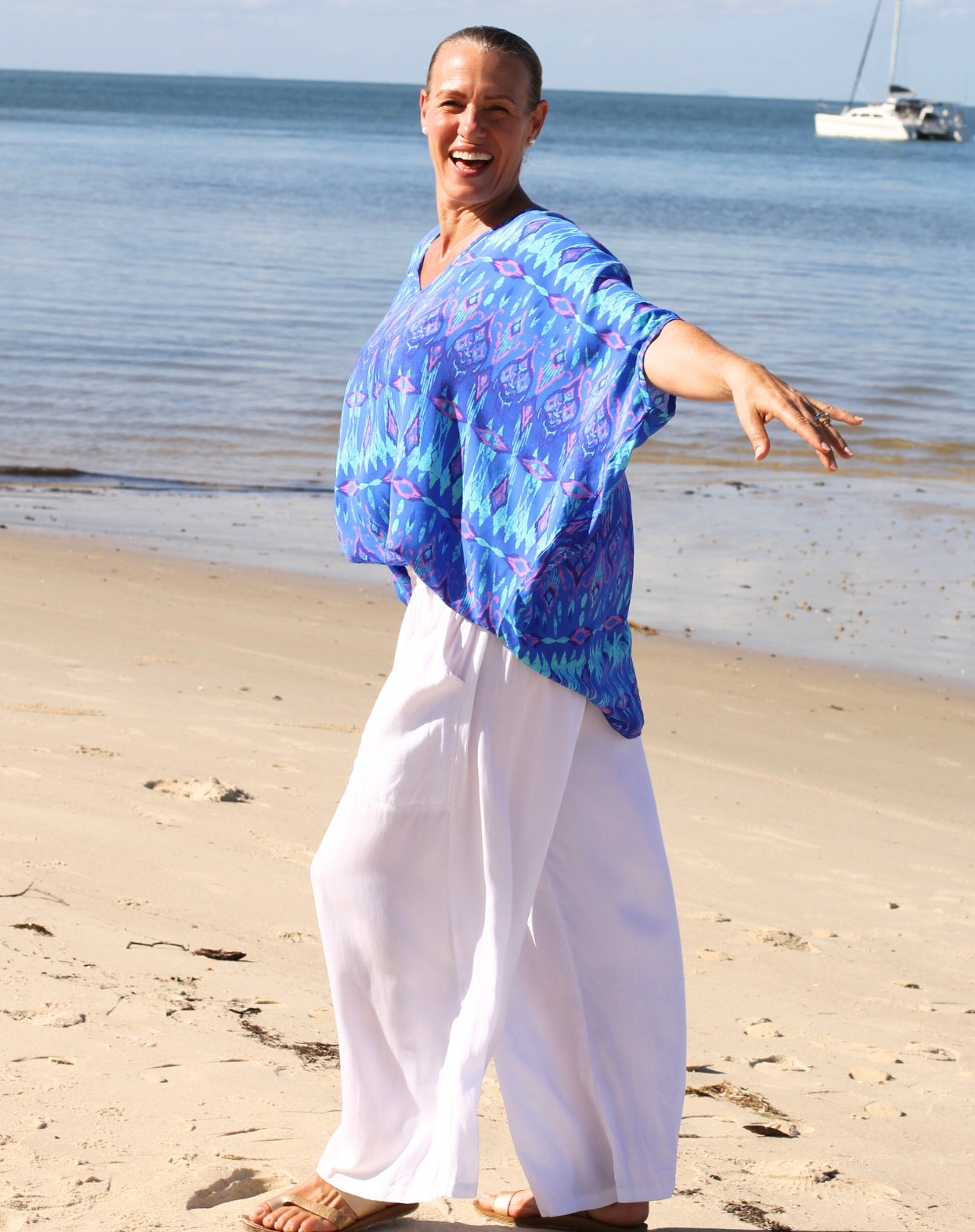 A smiling woman standing on a sandy beach with the ocean and a sailboat in the background, wearing a loose blue patterned top and white wide-leg pants.