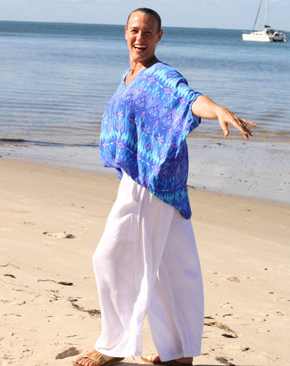 A smiling woman standing on a sandy beach with the ocean and a sailboat in the background, wearing a loose blue patterned top and white wide-leg pants.