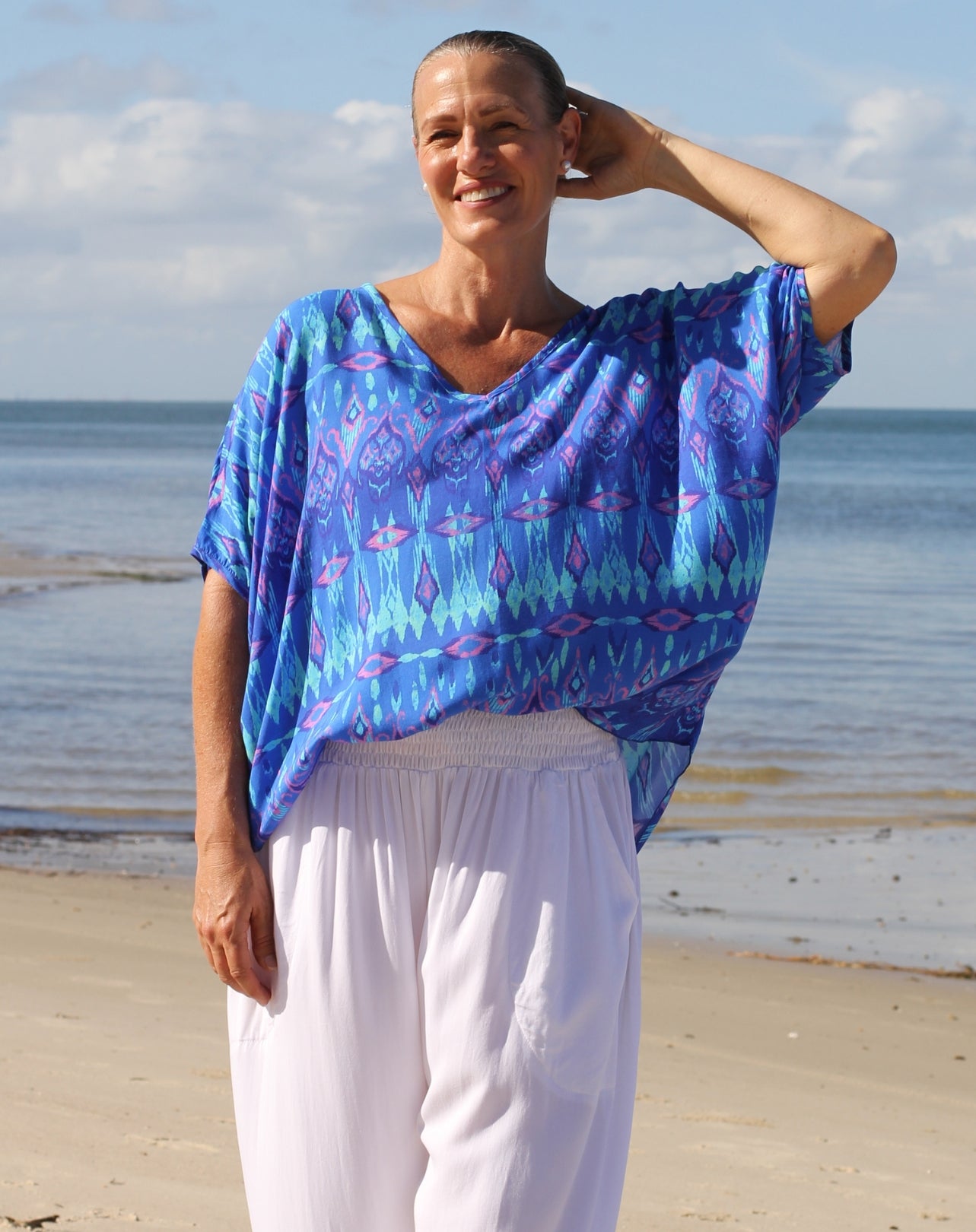 A woman standing on a sandy beach wearing a loose, short-sleeve V-neck top with a blue, purple, and teal geometric pattern, paired with white pants.