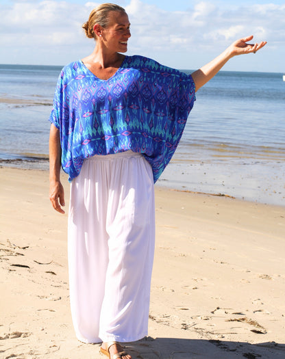 A woman standing on a sandy beach wearing a loose batwing style top in blue and purple patterned ikat top with white wide-leg pants, with the ocean and sky in the background.