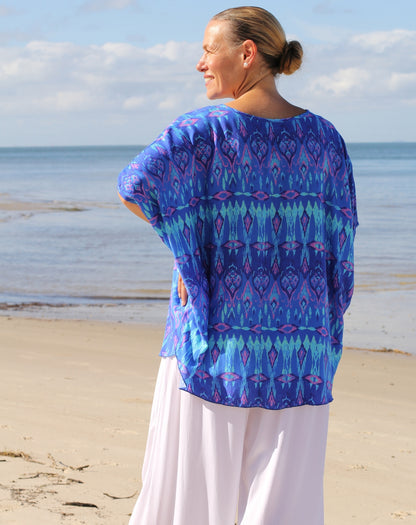 A woman standing on a sandy beach with the ocean and sky in the background, wearing a loose blue patterned ikat top and white pants, viewed from behind.