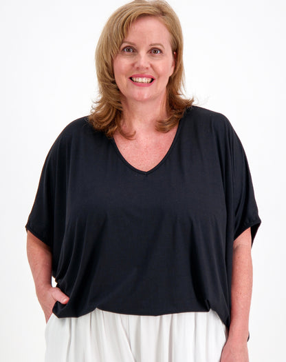 A smiling woman with blonde hair wearing a loose, short-sleeve V-neck top in black and white pants, standing against a plain white background.