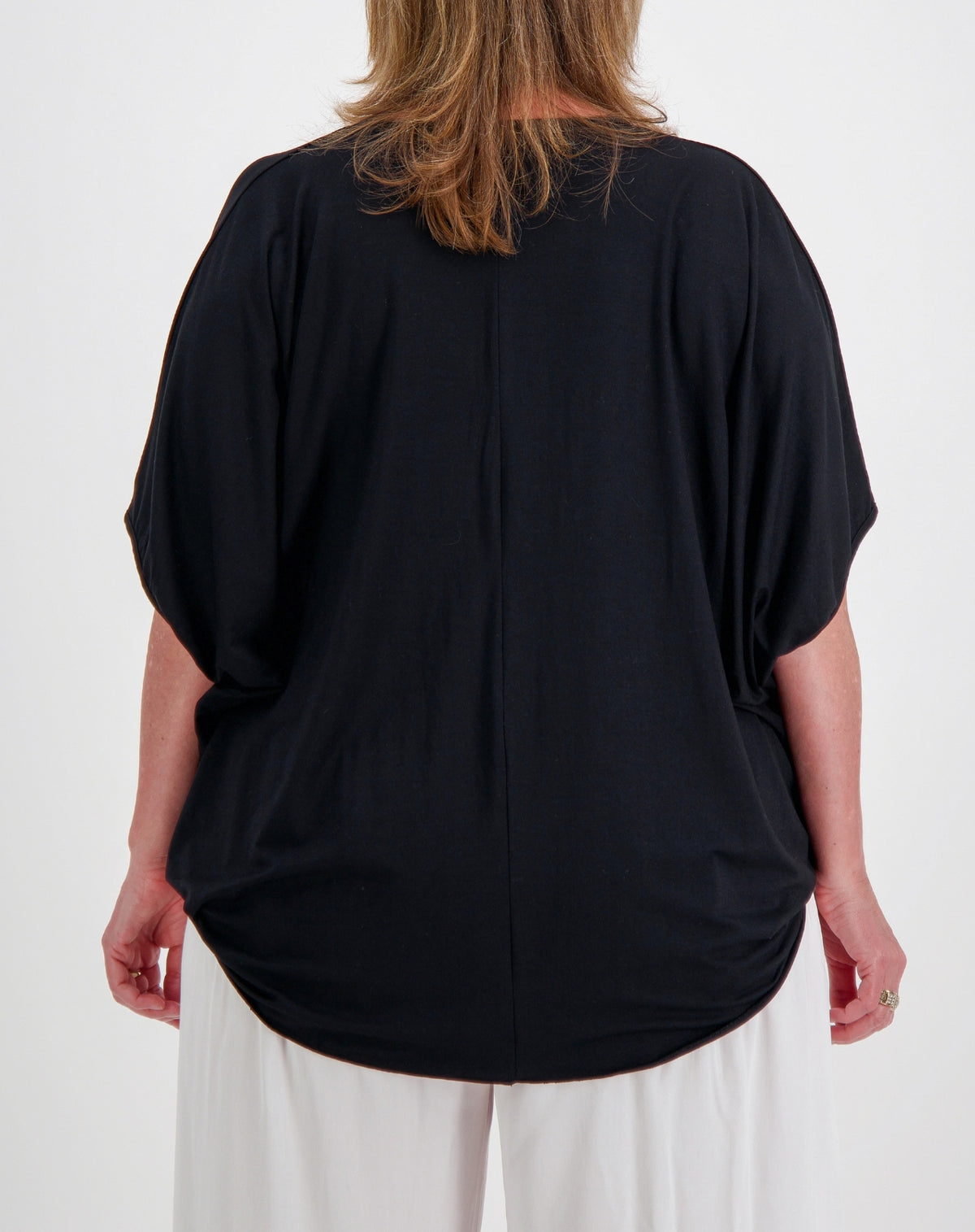 A woman viewed from the back wearing a loose black short-sleeve top and white pants against a plain white studio background.