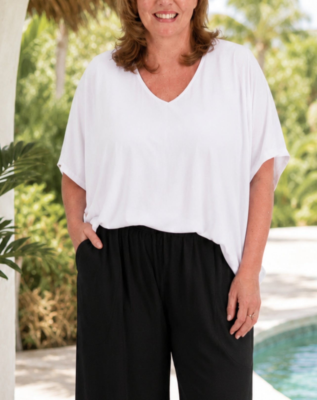 A woman wearing a white short-sleeve V-neck top and black pants, standing outdoors near a pool with green foliage in the background.