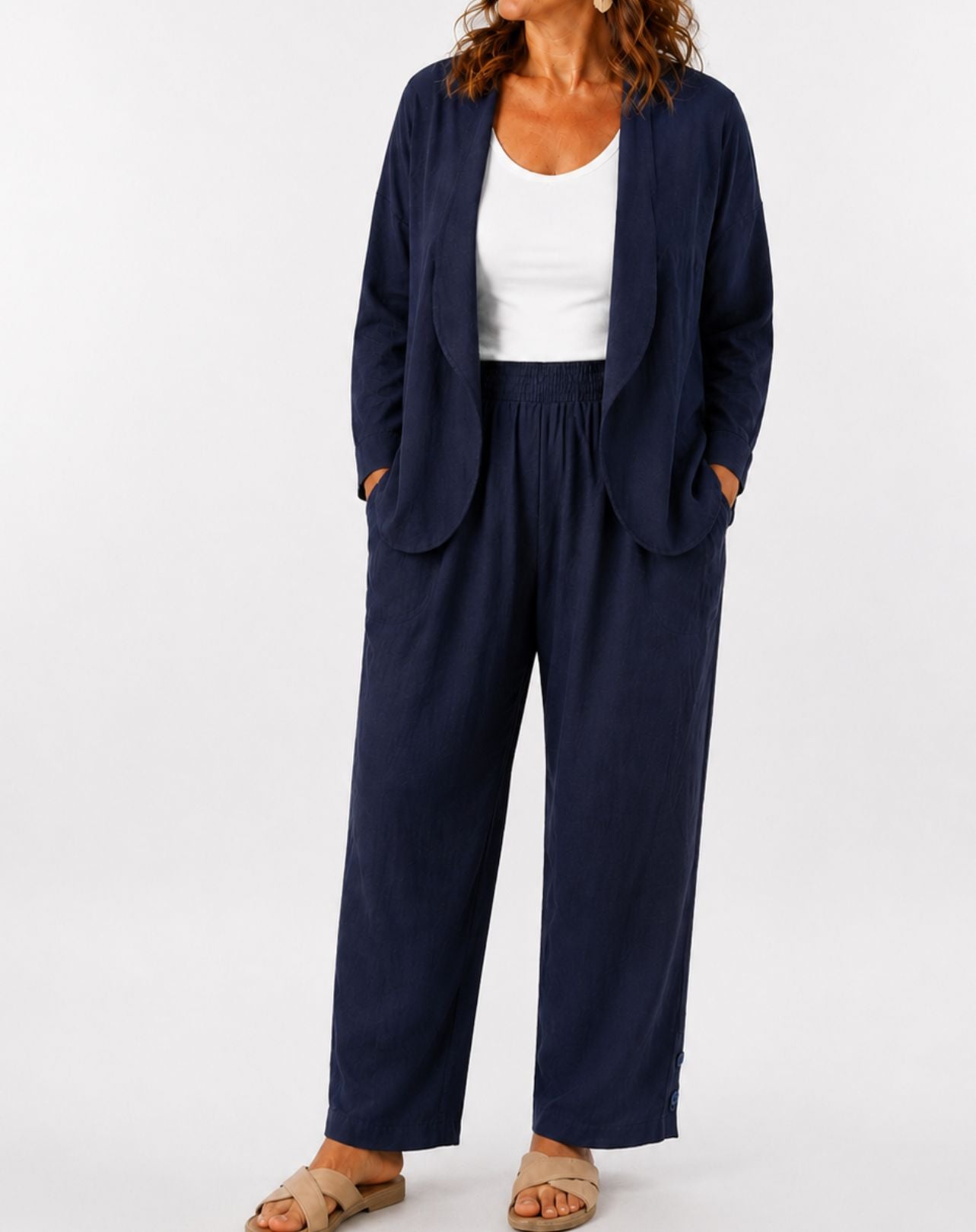 A woman wearing a navy blue two-piece linen look set consisting of a cropped open-front blazer and matching straight-leg trousers, styled with a white scoop tee and beige flat sandals against a plain white studio background.