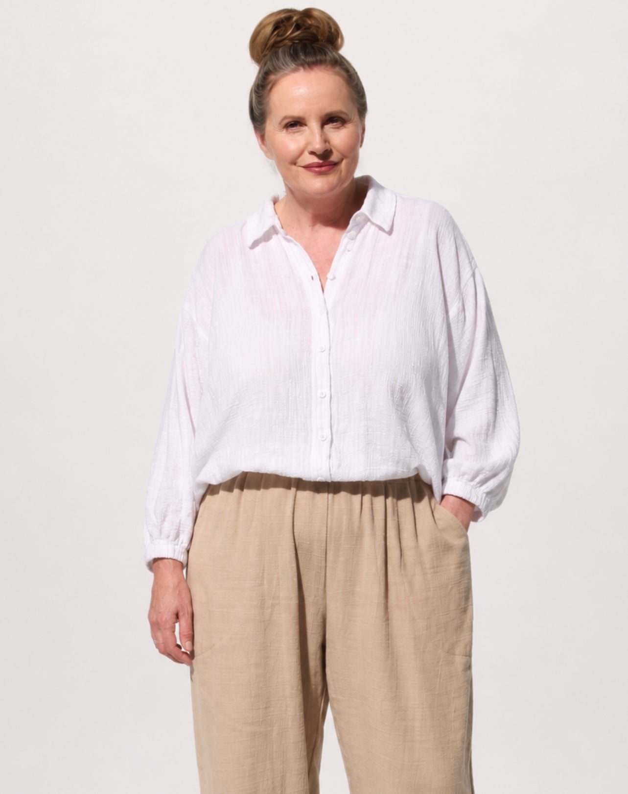 A woman modelling a white cotton comfort shirt long-sleeve button-up shirt with a relaxed, loose fit and beige linen trousers, standing against a solid off-white background.